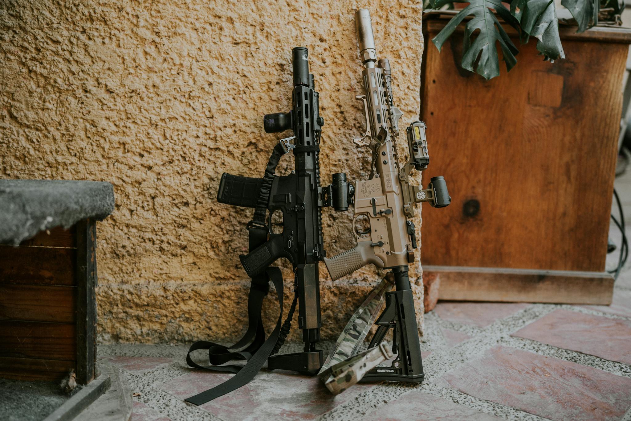 Two tactical rifles leaning against a rustic wall, showcasing military equipment.